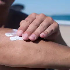 Dłoń rozsmarowuje krem z filtrem, zawierający m.in. diethylamino hydroxybenzoyl hexyl benzoate, na ramieniu na plaży.