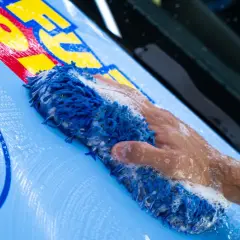 Mano con spugna blu insaponata mentre si lava un'auto con adesivi gialli e rossi.