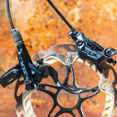 Shimano SLX M7120 hydraulic disc brakes, ready for any trail. The dark blue calipers and rotors are mounted on a rusty, textured background.