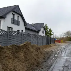 Nowy, szary płot dzieli posesje, sugerując konflikt z sąsiadem o ogrodzenie. Błotnista droga i sterta ziemi podkreślają napięcie.