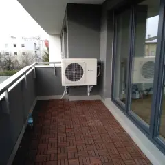 Klimatyzacja w bloku zamontowana na balkonie. Jednostka zewnętrzna na szarym tle, obok drzwi balkonowych.