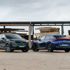 Dwa elektryczne SUV-y, Nissan Ariya i Kia EV6, stoją obok siebie na tle składu drewna.