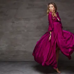 Kobieta w zwiewnej sukni w kolorze amarant, wiruje na tle szarej ściany.