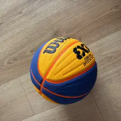 Piłka do koszykówki 3x3 Wilson z pomarańczowo-niebieskimi panelami leży na drewnianej podłodze.