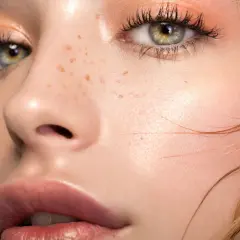 Occhi verdi con un delicato trucco lilla e pesca, labbra piene e lentiggini naturali.