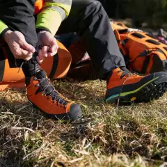 A person is lacing up bright orange hiking boots with a heel lock lacing system, preparing for an adventure with an orange backpack nearby.
