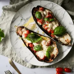 Dwa połówki bakłażana pieczonego z pomidorkami i mozzarellą, ozdobione listkami bazylii.