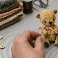 Mały, ręcznie robiony miś z materiału, ozdabiany wstążką, obok stosu tkanin i akcesoriów.