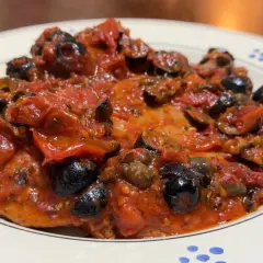Bistecche alla pizzaiola, un piatto succulento con pomodoro, olive nere e capperi, servito su un piatto bianco con decorazioni blu.