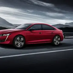 Voiture rouge Peugeot 508, un modèle à éviter, roulant sur une route avec des montagnes en arrière-plan sous un ciel dramatique.