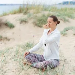 Kobieta siedzi na plaży, praktykując pranajamę. Jej oczy są zamknięte, a palce wskazujące dotykają nosa, kontrolując oddech.