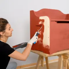 Mujer con guantes blancos pinta con rodillo una caja de madera de color terracota.