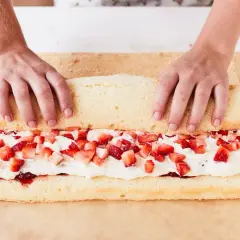 Hände rollen einen Biskuitteig mit Sahne und Erdbeeren für ein Erdbeerrolle Rezept auf.