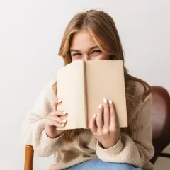 Dziewczyna z uśmiechem ukrywa twarz za książką, zanurzona w świecie literatury young adult.