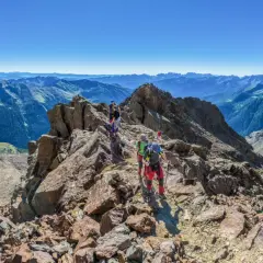 Grupa ludzi wyrusza na trekking po skalistym szczycie, podziwiając rozległe pasmo górskie w oddali.