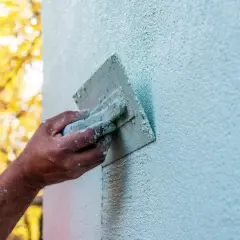 Ręka zacierająca tynk silikonowy na jasnoniebieskiej ścianie. W tle jesienne drzewa w słońcu.