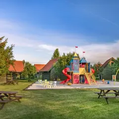 Kolorowy plac zabaw w kształcie zamku, huśtawki i stoły piknikowe na trawniku. W tle drewniane domki, idealne na łebska chata.