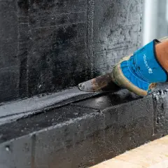 Rękawica z narzędziem nakłada czarną masę izolacyjną, chroniąc fundamenty przed wilgocią.