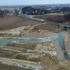 Budowa kanału wodnego w deszcz nawalny. Widać koparki, ciężarówki i umocnione brzegi. W tle droga i zabudowania.