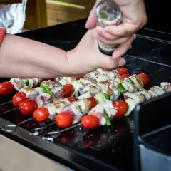 Brochettes de viande, tomates et courgettes sur une plancha à gaz. Une main assaisonne les brochettes.