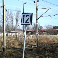 Wskaźnik W24 z numerem 12 przy torach kolejowych. W tle budynki i drzewa.