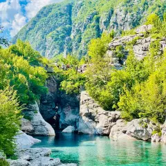 Aguas turquesas fluyen entre rocas y vegetación exuberante, bajo imponentes montañas en Albania. Un puente peatonal cruza el cañón, invitando a explorar las **montañas malditas de Albania**.