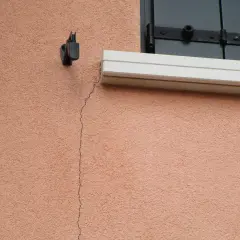 Una crepa sottile attraversa l'intonaco rosa di un muro esterno, vicino a un supporto metallico e a una finestra con persiana. Utile per capire come riempire le crepe nei muri esterni.