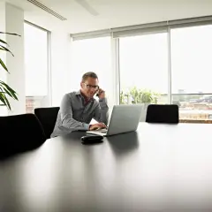 Biznesmen rozmawia przez telefon, pracując na laptopie. W tle okna z widokiem na miasto, a obok rośliny. To jego koszt uzyskania przychodu.