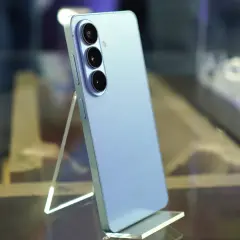 Jasnoniebieski Samsung Galaxy S26 z potrójnym aparatem z tyłu, stojący na przezroczystej podstawce.