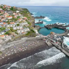 Urokliwe miasteczko na Maderze z kolorowymi domkami na zboczu klifu, czarną plażą i naturalnymi basenami w oceanie.