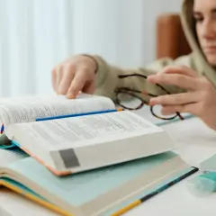 Chłopak w bluzie pochyla się nad książkami, analizując **motywy literackie**. Na stole leżą otwarte tomy, okulary i kolorowe karteczki.