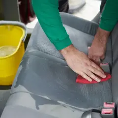 Ręce czyszczą siedzenie samochodowe czerwoną gąbką, obok żółte wiadro z wodą. Praktyczny poradnik, jak usunąć plamy z tapicerki samochodowej.