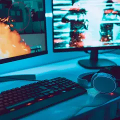 Gaming-Setup mit zwei Monitoren, Tastatur und Kopfhörern. Auf dem linken Bildschirm lodert Feuer, rechts sind Charaktere zu sehen. Perfekt für VA oder IPS.
