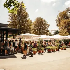 Sonniger Biergarten im Schlossgarten mit vielen Gästen unter schattenspendenden Schirmen.