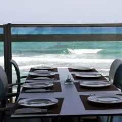 Mesa puesta en el chiringuito la sal, con platos y cubiertos, frente a un mar turquesa y olas rompiendo.