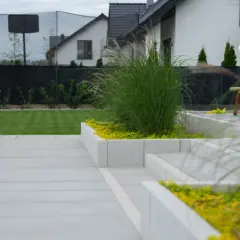 Nowoczesny taras z betonowych płyt i podniesionych rabat. Inspiracje dla ogrodu z trawami ozdobnymi i żółtą roślinnością.