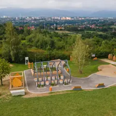 Nowoczesny plac zabaw i siłownia plenerowa, otoczone zielenią, z widokiem na miasto. W oddali widać trzy lipki.