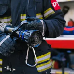 Strażak w rękawicach trzyma prądownicę. Zastanawiasz się, jakie są stopnie w PSP po ilu latach?