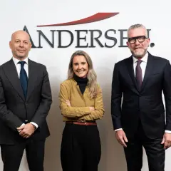 Tres personas posan frente al logo de Andersen. El hombre de la izquierda, calvo, viste traje gris. La mujer del centro, con brazos cruzados, lleva una blusa ocre. El hombre de la derecha, con gafas, viste traje oscuro. Un momento clave, como un ejempl...