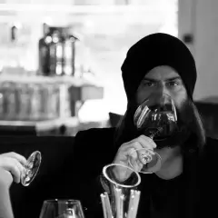 Ein Mann mit Bart und Mütze trinkt Wein. Er hält das Glas am Stiel, eine elegante Art, wie trinkt man wein.