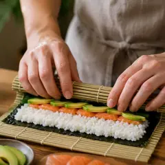 Ręce zwijają sushi z łososiem i awokado na macie bambusowej. Obok butelka octu ryżowego do sushi.