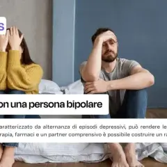 A couple sits apart on a bed, one covering their face, the other holding their head.