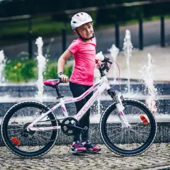 Uśmiechnięta dziewczynka w kasku stoi obok swojego białego roweru dla 7 latki, z fontannami w tle.