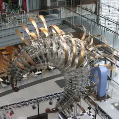 Duża, metalowa rzeźba przypominająca szkielet dinozaura w centrum handlowym.