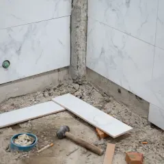 Début des travaux pour carreler une salle de bain : des carreaux de marbre blanc sont posés sur le sol, près d'un marteau et d'un seau.