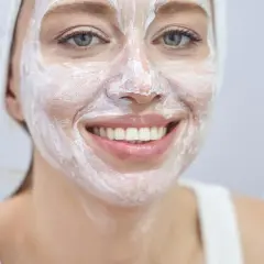 Lächelnde Frau mit weißer Gesichtsmaske und Handtuch auf dem Kopf. Wie oft Maske Gesicht? Regelmäßige Pflege für strahlende Haut.