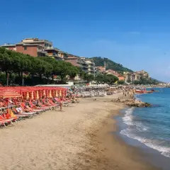 Plage d'Imperia animée, avec des rangées de parasols rouges et des chaises longues sur le sable. La mer bleue étincelle sous un ciel clair.