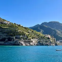 A scenic view from a Naples to Amalfi Coast ferry, showing terraced hillsides dotted with houses, a grand villa, and the sparkling blue sea.