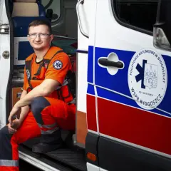 Mężczyzna w pomarańczowym stroju ratownika medycznego siedzi w otwartych drzwiach karetki.