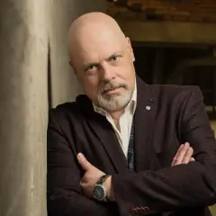 Marek Krajewski w eleganckim stroju, oparty o ścianę, zamyślony. W tle industrialne wnętrze, sugerujące jego życie prywatne.